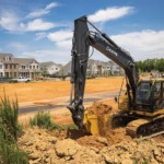 Una Excavadora 160P-Tier mueve tierra en un lugar de trabajo con un desarrollo habitacional en el fondo.