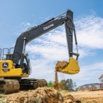 Una Excavadora 200G mueve tierra en un lugar de trabajo con un desarrollo habitacional en el fondo.